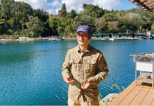 事務所のデッキから舞根湾を背景に、地域再生の想いを語る副理事長 畠山信さんの写真